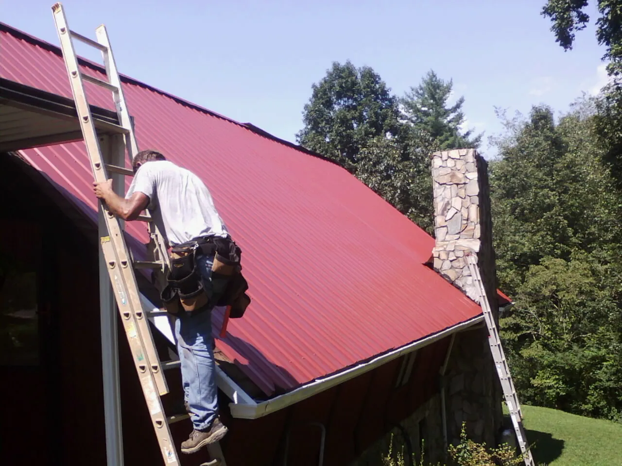 Roofer on a ladder beside a red metal roof and stone chimney — Soffit & Fascia Repair by Roofing Brothers in Castleton